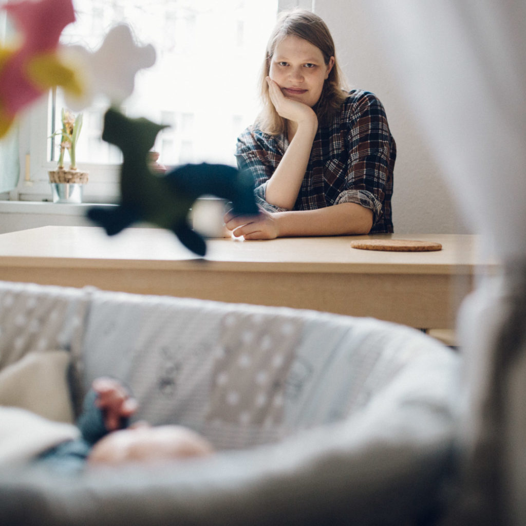 Neugeborenenfotos Berlin Familienreportage Berlin-Wedding: Baby liegt im Vordergrund in Stubenwagen, Mutter sitzt im Hintergrund damit Tisch