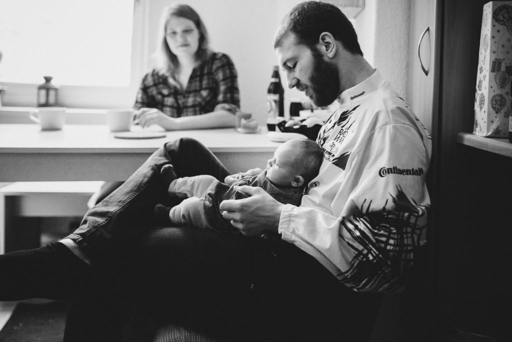 Neugeborenenfotos Berlin: Vater sitzt mit Baby in Küche. Mutter im Hintergrund bei Familienreportage in Berlin Wedding