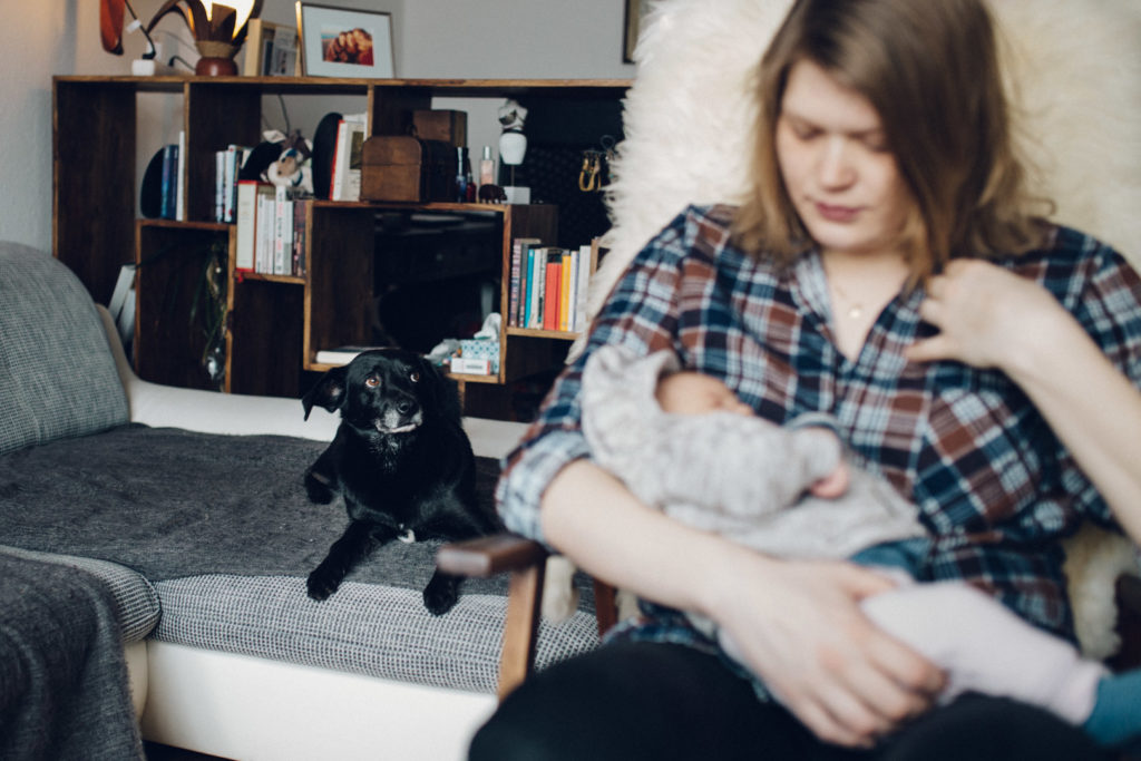 Neugeborenenfotos Berlin Familienreportage Berlin-Wedding: Mutter mit Neugeborenem auf dem Arm. Hund im Hintergrund auf dem Sofa
