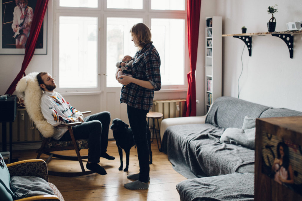 Neugeborenenfotos Berlin Familienreportage Berlin-Wedding: Vater sitzt erschöpft auf Schaukelstuhl. Mutter mit Baby auf dem Arm steht davor
