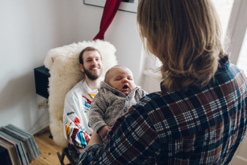 Neugeborenenfotos Berlin: Baby gähnt. Lächelnder Vater im Hintergrund bei Familienreportage in Berlin-Wedding