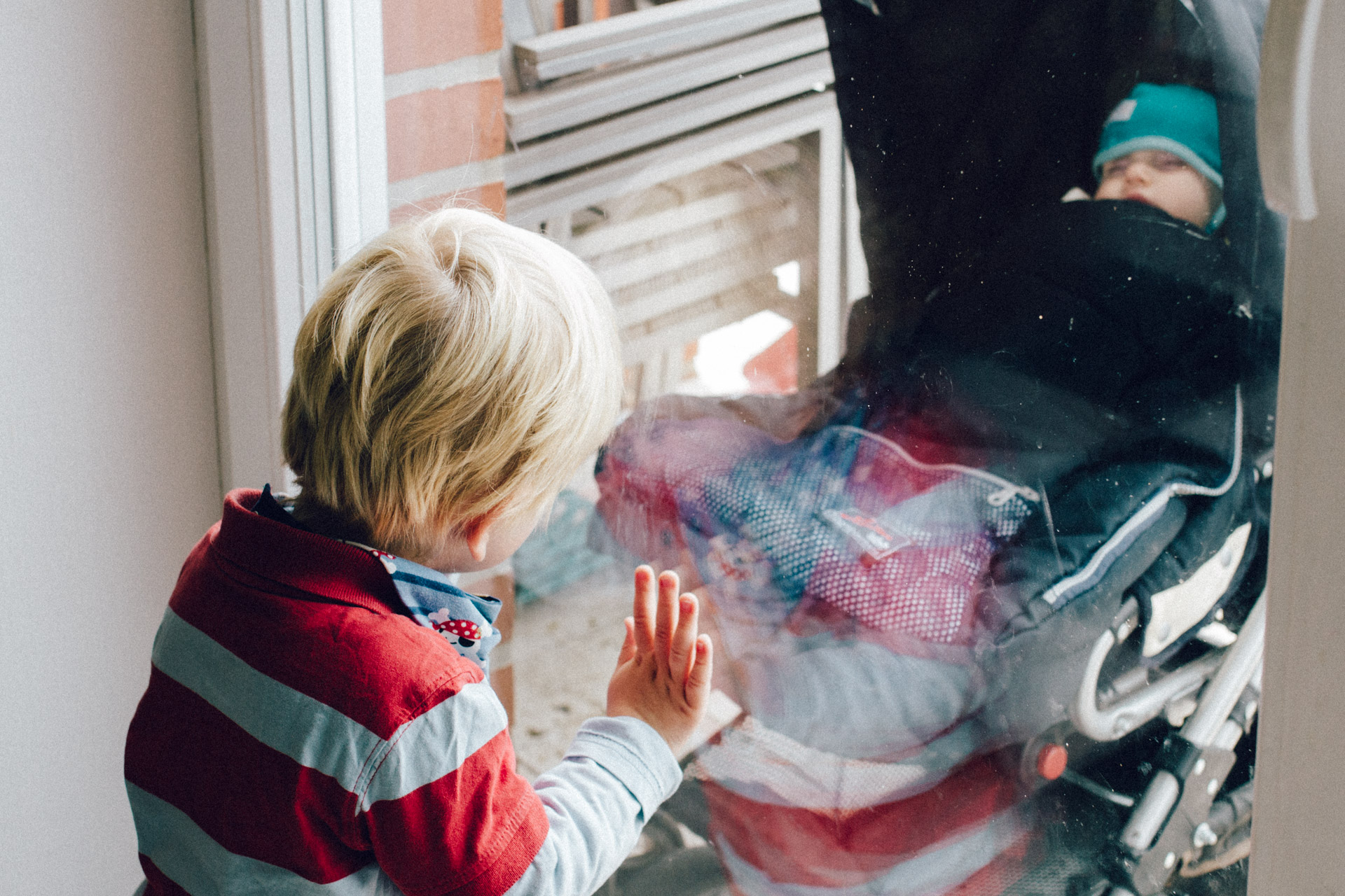 Familienreportage Berlin: blonder Junge in rot gestreiftem Pullover schaut aus dem Fenster vor dem ein Baby im Kinderwagen schläft