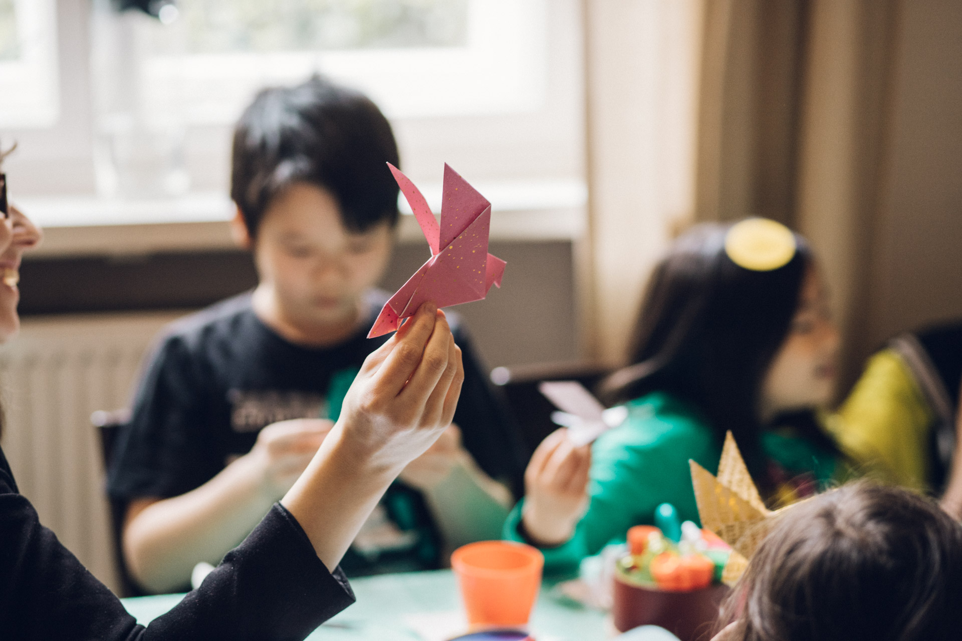 Familienreportage Berlin: Kinder basteln im Hingrund. Frau hält Origami Vogel im Vordergrund hoch